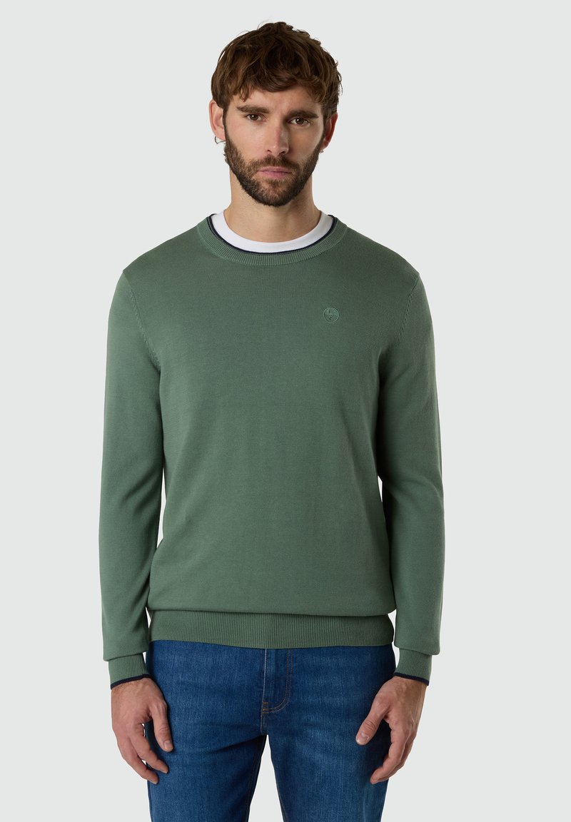 Uomo con barba che indossa un maglione verde girocollo sopra una camicia bianca e jeans blu, in piedi contro uno sfondo chiaro semplice.