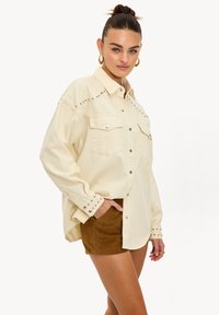 Camisa abotonada de color beige claro con acentos de tachuelas en los hombros y en los puños, con dos bolsillos en el pecho y un ajuste relajado. Combinada con pantalones cortos marrones.