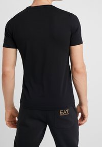 Musta lyhythihainen T-paita, valmistettu sileästä kankaasta. Tiimalasimallinen leikkaus ja pyöreä pääntie. Ominaisuuksina olkapään läheisyydessä oleva logokoriste.