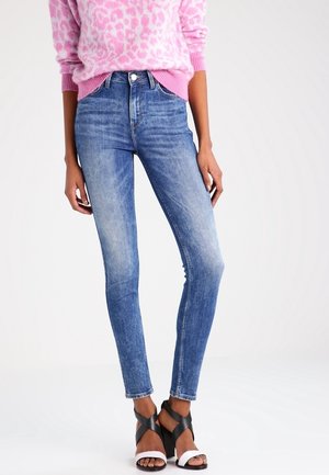 Model iført højtaljede skinny blå jeans, lyserød leopardprint trøje og sorte og hvide remsandaletter med høj hæl på hvid baggrund.