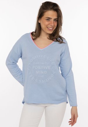 Frau lächelnd, trägt einen hellblauen Pullover mit dem Text "Positive Life Positive Vibes Positive Mind Club" und weiße Hose, steht vor einem weißen Hintergrund.