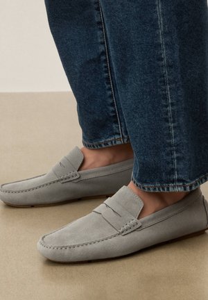 Világosszürke velúr mokaszinok sötétkék farmernadrággal viselve, részletes varrással és sima talppal, bézs felületen.