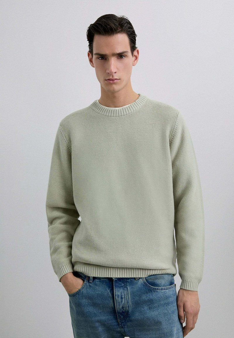 Suéter de punto verde claro con cuello redondo y puños acanalados. Se lleva con jeans azules desgastados, que presentan un corte relajado y un diseño clásico.