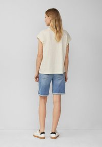 Lichtbeige overhemd met korte mouwen, denim korte broek met opgerolde mouwen en witte sneakers met zwarte accenten, gezien vanuit de achterkant.