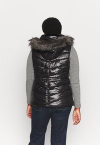 Schwarze Steppweste mit glänzender Oberfläche, ausgestattet mit einer Kapuze mit Pelzbesatz, gestepptem Design und seitlichen Taschen, getragen über einem grauen Langarmoberteil.