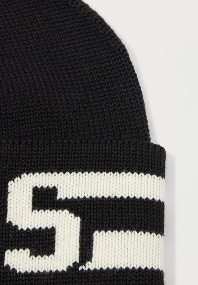Gorro de punto negro con puño doblado, que presenta franjas y texto blancos en negrita. El material parece suave y texturizado, adecuado para el clima frío.