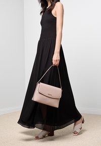 Kobieta w czarnej sukni maxi bez rękawów, trzymająca jasnoróżową torebkę i nosząca wzorzyste sandały na obcasie, stojąca na beżowym dywanie.