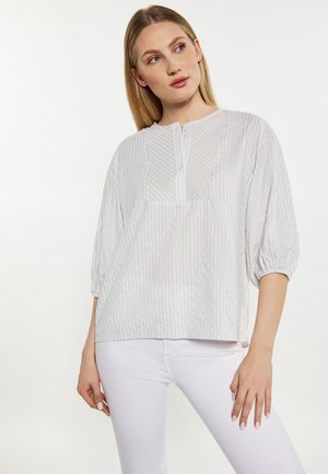 Femme portant un chemisier ample à rayures bleu clair et blanc avec des manches trois-quarts et un pantalon blanc ajusté sur un fond uni.