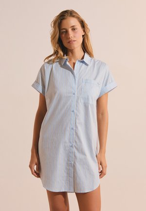 Mujer con un vestido camisa de manga corta y botones, con rayas verticales azules y blancas, de pie frente a un fondo beige liso.