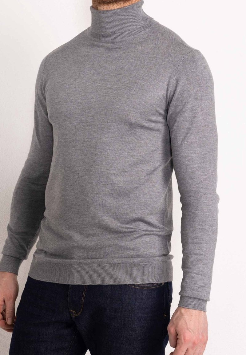 Pull à col roulé gris en tissu doux, avec manches longues, poignets côtelés et coupe slim. Porté avec un jean foncé.
