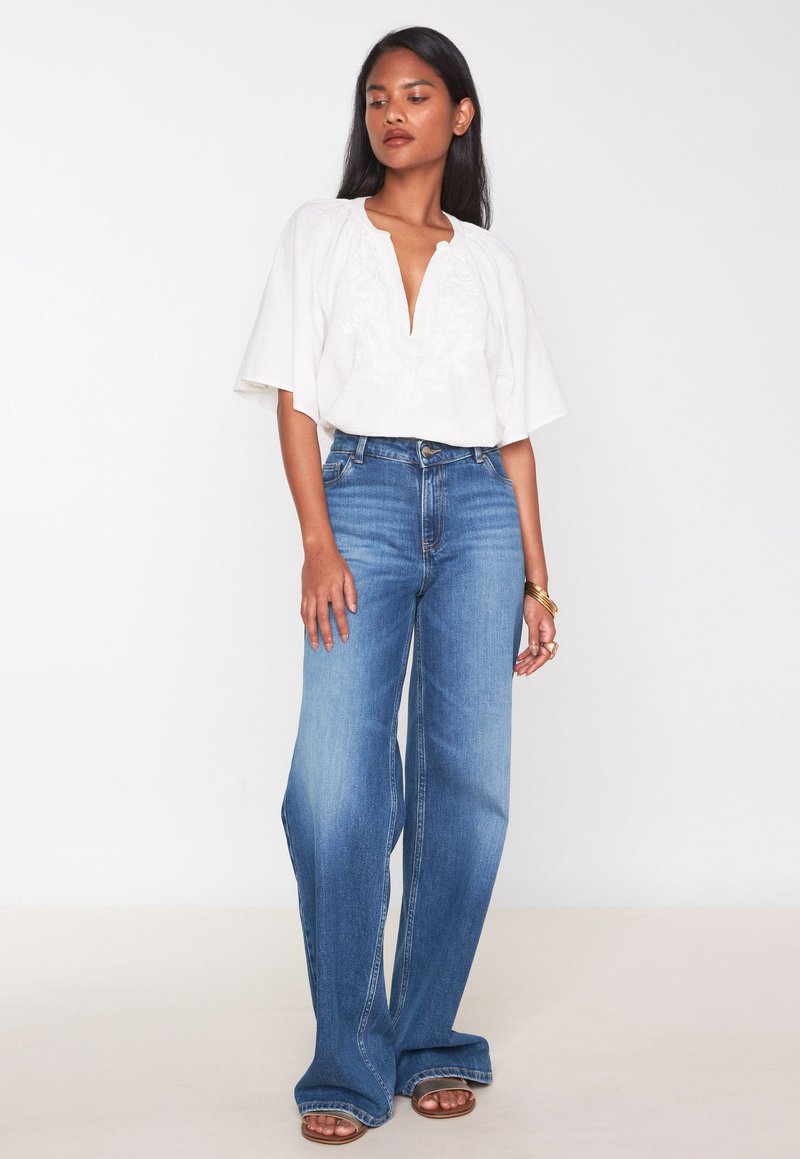 Witte, gestructureerde blouse met een V-hals en uitlopende mouwen, gecombineerd met een wijde blauwe jeans. Sandalen met een kleine hak maken de outfit compleet.