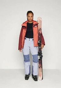 Στολή σκι: πορτοκαλί-κόκκινο μπουφάν με μαύρες λεπτομέρειες, εφαρμοστό μαύρο μπλουζάκι, ανοιχτό μπλε παντελόνι σκι με ναυτικές ραφές, μαύρες μπότες σκι, κρατώντας σκι.