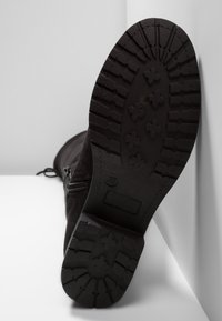 Suela de bota negra con un patrón de surcos profundos apoyada verticalmente contra una superficie blanca, exhibiendo la talla 37 estampada en la suela.