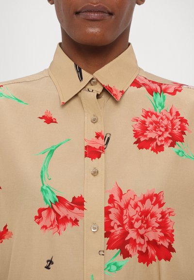 Camisa beige con gran estampado de flores rojas. Cuenta con cuello, cierre de botones al frente y sutiles acentos verdes. Textura de tela suave.