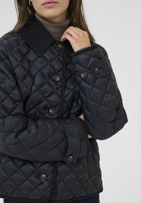 Black quilted jacket with a diamond pattern, featuring a corduroy collar, oversized buttons, and textured fabric.