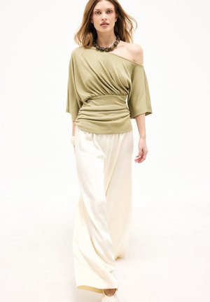 Žena kráča, oblečená v olivovom top s odhalenými ramenami a záhybmi, široké biele nohavice a masívny náhrdelník z korálikov na bielom pozadí.