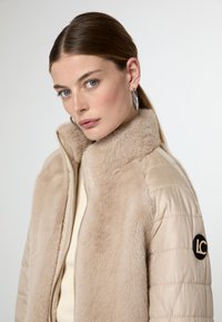 Giacca bomber beige con colletto in pelliccia sintetica, chiusura con zip e texture trapuntata. Presenta un logo circolare sulla manica sinistra.