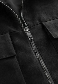 Schwarze Wildlederjacke mit einem silbernen Metallreißverschluss, zwei Fronttaschen und einer glatten Textur, die die Nähte und Designdetails hervorhebt.