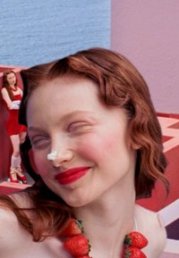 Nahaufnahme einer Frau mit welligem roten Haar, die eine Erdbeer-Halskette und leuchtend roten Lippenstift trägt, mit einem verspielten Ausdruck und einer cremigen Rückstände auf ihrer Nase.