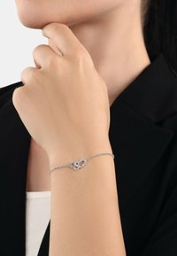 Bracciale d'argento con design di cuori intrecciati, caratterizzato da piccole pietre scintillanti. Le catene sono delicate e lucide su uno sfondo neutro.