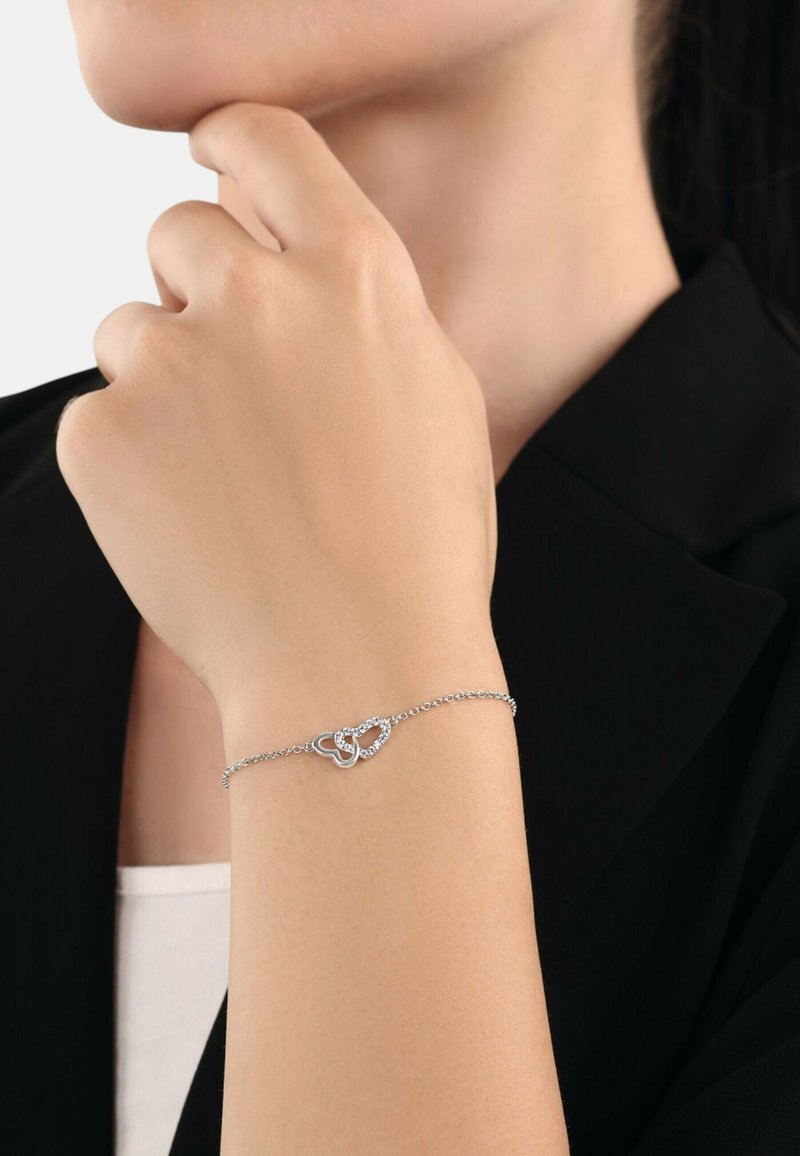 Bracciale d'argento con design di cuori intrecciati, caratterizzato da piccole pietre scintillanti. Le catene sono delicate e lucide su uno sfondo neutro.