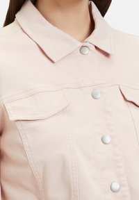 Světle růžová džínová bunda s knoflíkovým zapínáním, stříbrnými doplňky, dvěma náprsními kapsami a klasickým límcem s viditelnými švy.