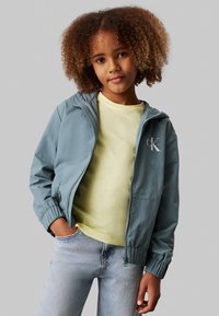 Lys blå vindjakke med hætte, med elastiske manchetter, båret over en bleg gul skjorte og lyse denimjeans. Logo på brystet.