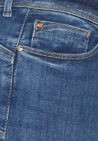 Jeans de ganga em lavagem azul escura com costura visível. Apresenta um pequeno bolso frontal, acessórios em bronze e uma textura suave.