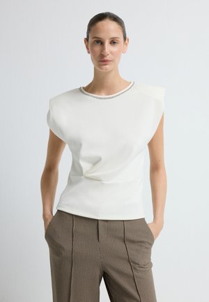 Biała bluzka z wyraźnie zarysowanymi ramionami, z twistem z przodu i ozdobnym dekoltem. Połączona z brązowymi spodniami w kratę.