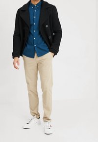 Czarny płaszcz typu peacoat, niebieska koszula z guzikami, beżowe spodnie, białe trampki. Płaszcz ma podwójne guziki i szerokie klapy. Teksturowana tkanina.