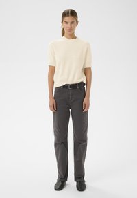 Kremasti kratki pleteni pulover s kratkim rukavima, u kombinaciji s tamnosivim hlačama ravnog kroja i crnim cipelama, stoji protiv neutralne pozadine.