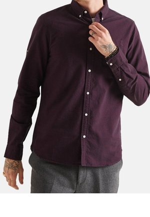 Hombre con una camisa de botones de color púrpura oscuro y pantalones grises, ajustándose el cuello, mostrando tatuajes en las manos y pulseras en las muñecas.
