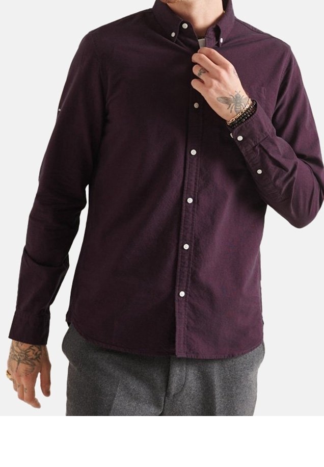 Uomo che indossa una camicia button-up viola scuro e pantaloni grigi, mentre regola il colletto, mostrando tatuaggi sulle mani e braccialetti ai polsi.