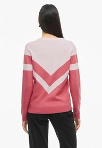 Rožinė ir šviesiai rožinė megztinis su V formos dizainu nugaroje, ilgomis rankovėmis ir raukšlėmis ant rankovių, derinamas su juodomis kelnėmis.