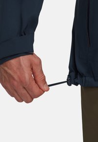 Veste bleu marine avec une fermeture à cordon au niveau des poignets. Une main tire sur le cordon, mettant en valeur la texture du tissu et les détails de design.