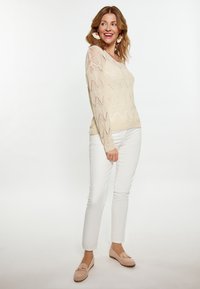 Strikket cremefarvet sweater med zigzag mønster, parret med hvide tætsiddende bukser og lyserøde loafers. Modellen har bølget hår og store øreringe.