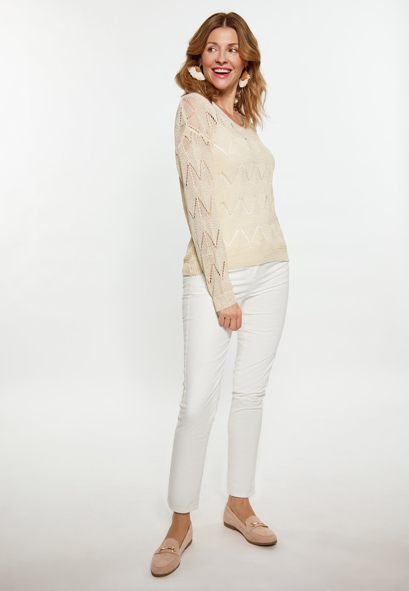 Strikket cremefarvet sweater med zigzag mønster, parret med hvide tætsiddende bukser og lyserøde loafers. Modellen har bølget hår og store øreringe.
