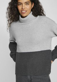 Suéter de punto en gris claro y gris oscuro con cuello alto, que presenta una textura acanalada y un diseño de bloques de color contrastantes en las mangas.