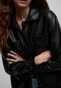 Black leather shirt with a collared neckline, featuring two front pockets, button closures, and a smooth texture.