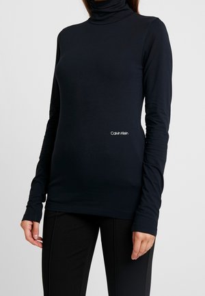 Frau trägt ein eng anliegendes schwarzes Langarm-Top mit Rollkragen und Calvin Klein Logo oberhalb der Taille, kombiniert mit schwarzen Hosen.