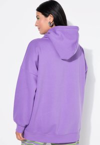 Fioletowa bluza z kapturem i sznurkiem do ściągania, w oversize'owym kroju, z prążkowanymi mankietami i dołem. Gładka tekstura, bez widocznych wzorów ani grafik.