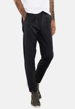 Schwarze, taillierte Hose mit gefaltetem Vorderteil und seitlichen Taschen. Hergestellt aus einem weichen, strukturierten Stoff. Kombiniert mit grauen Sneakern.