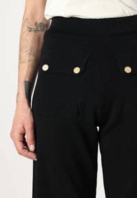 Svarte bukser med en tilpasset midje, som har to frontlommer med gullknapper. Mykt, teksturert stoff komplementerer designet.