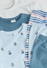 Trois bodies pour bébé en blanc, bleu foncé et bleu clair avec des motifs de voiliers, assortis à des pantalons à taille élastique rayés et unis bleus.