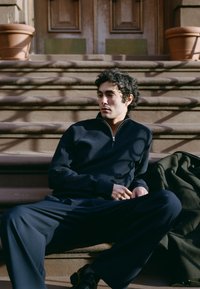 Mężczyzna o kręconych włosach w ciemnym ubraniu siedzący na zewnętrznych kamiennych schodach obok zielonego płaszcza w miękkim świetle słonecznym.