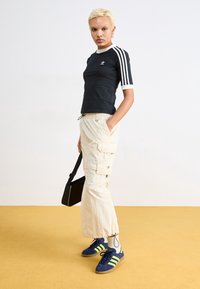 Černá krátká košile s bílými pruhy, světle béžové cargo kalhoty s kapsami a tmavě modré boty se žlutými detaily. Černá taška přes rameno.