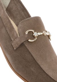 Mocassino in suede marrone con punta arrotondata, accentato da hardware dorato e una fascia decorativa con strass sul lato. Fodera interna liscia.