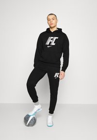 Zwarte hoodie en bijpassende broek met wit "FC" logo, staande op een grijze voetbal, accessorized met witte en blauwe schoenen.