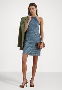 Teal värinen halterneck-mekko, jossa on geometrinen kuvio ja muodostaa kimalteita. Yhdistetty oliivinvihreään takkiin ja ruskeaan laukun, korostaen modernia ilmettä.