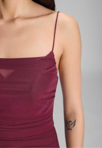 Bordeauxfarbenes ärmelloses Oberteil mit taillierter Passform, das aus weichem, drapiertem Stoff und dünnen Trägern besteht, akzentuiert durch ein Tattoo am linken Arm.